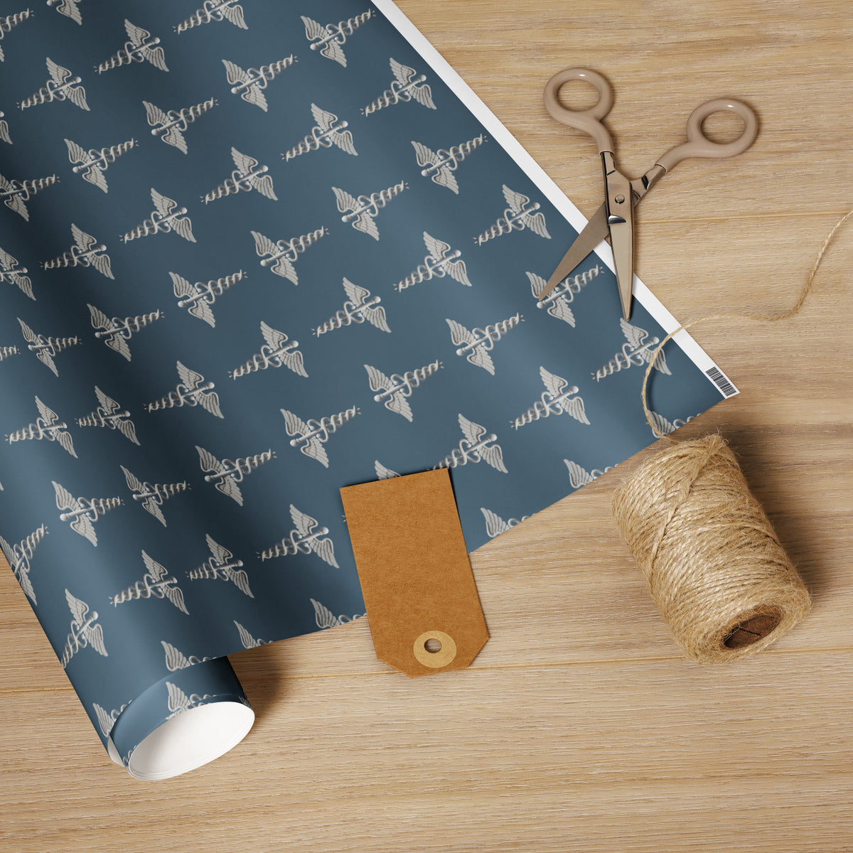 Medical Caduceus Wrapping Paper, white glossy finish: embroidered-look caduceus pattern on an ocean-toned ground, unrolled on a wooden table with scissors, twine and a kraft gift tag